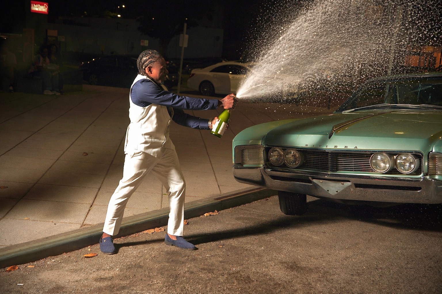 Man in a cream suit spraying champagne beside a classic Buick Riviera at night.