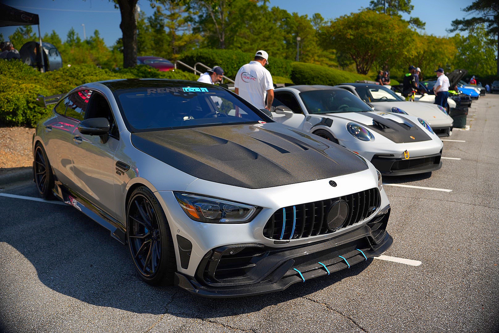 Silver Mercedes-AMG GT with carbon hood beside a lineup of Porsches.
